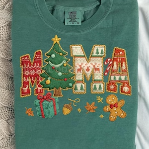 May include: A teal green sweatshirt with the word "MAMA" embroidered in festive Christmas designs. The "M"s feature reindeer and snowflake patterns, while the "A"s have Christmas trees and candy canes. A Christmas tree, gift, and gingerbread man are also embroidered.