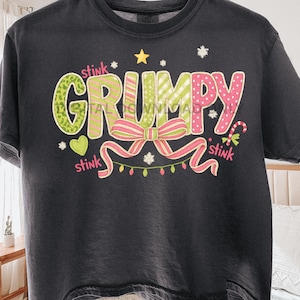 Op de afbeelding: Donkergrijs t-shirt met het woord "GRUMPY" in groen en roze, versierd met een strik, sterren en het woord "stink". Het shirt heeft een ronde hals en korte mouwen.