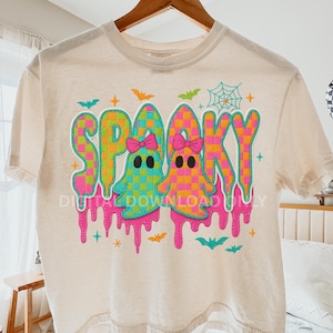 Può includere: T-shirt color crema con grafica colorata "Spooky". Il design presenta due figure di fantasmi con fiocchi, circondate da pipistrelli, stelle e una ragnatela. La parola "Spooky" è in un motivo a scacchi con effetto gocciolante.