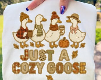 Op de afbeelding: Wit sweatshirt met een geborduurd ontwerp van vier ganzen in herfstoutfits, met koffiebekers en een pompoen. De tekst "JUST A COZY GOOSE" staat eronder. Alleen digitale download.