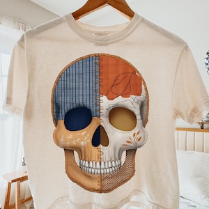 Peut inclure: T-shirt crème avec un motif de crâne coloré. Le crâne est divisé en sections avec divers motifs, notamment du carreau bleu, de l'orange et des motifs floraux. Le motif est centré sur le t-shirt.