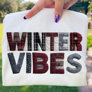 May include: A white sweatshirt featuring the text "WINTER VIBES" in appliquéd fabric letters. The letters use a variety of patterns, including plaid and polka dots. The sweatshirt is held up by a hand with blue nail polish.
