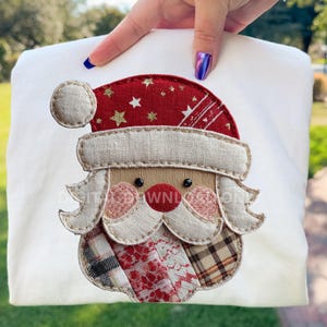 May include: A white fabric with an embroidered Santa Claus design. The Santa Claus has a red hat with gold stars, a beige beard, and a red nose. The design is made of various fabrics and patterns.