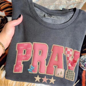 May include: Dark gray sweatshirt with the word "PRAY" appliqued in pink, with decorative elements including bows, stars, and a checkered pattern. The sweatshirt is folded, and a hand with a gold bracelet is visible.