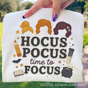 May include: White t-shirt with the phrase "Hocus Pocus time to FOCUS" in black and tan lettering. The design includes illustrations of three hairstyles, a pencil, a bat, stars, a book, a beaker, and a cauldron.