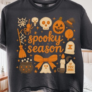 Pode incluir: T-shirt cinza escuro com um design de Halloween. O design inclui uma aranha, uma caveira, uma abóbora, um chapéu de bruxa e as palavras "spooky season". Também há um laço, um fantasma e o texto "IF YOU DARE."