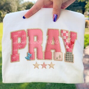 May include: White sweatshirt with the word "PRAY" in pink, embroidered with gold trim. The letters have various patterns, including bows, stripes, and a checkered design. Three gold stars are embroidered below the word.