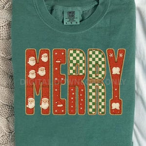 Könnte beinhalten: Ein teal-grünes Sweatshirt mit dem Wort "MERRY" in einem festlichen Design. Die Buchstaben sind rot umrandet und haben eine strukturierte Optik. Der Buchstabe "M" zeigt Santa-Gesichter, und der Buchstabe "E" ist ein grün-weißes Schachbrettmuster.