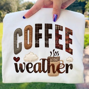 May include: White sweatshirt featuring the words "COFFEE weather" in brown, plaid, and tan appliqued letters. A coffee mug and clouds are also appliqued on the shirt. The sweatshirt is held up by a hand with blue nail polish.