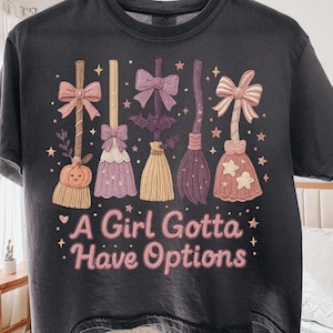 May include: Dark gray t-shirt featuring a whimsical design of five decorative witch brooms in various colors, including pink, purple, and gold. The text "A Girl Gotta Have Options" is printed below the brooms in pink and white. The design is surrounded by stars.