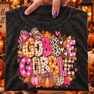 May include: A black mineral-washed t-shirt featuring a colorful graphic with the text GOBBLE GOBBLE. The design includes stylized letters in orange, pink, and brown patterns, decorated with a large bow, autumn leaves, and three pumpkins.