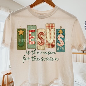 Könnte beinhalten: Cremefarbenes T-Shirt mit dem Aufdruck "JESUS is the reason for the season" auf der Vorderseite. Das Wort "JESUS" ist in einem dekorativen Stil gehalten, wobei jeder Buchstabe auf einem anderen rechteckigen Patch steht. Die Patches sind grün, rot und blau.