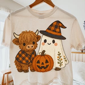 以下が含まれることがあります： ハロウィーンをテーマにしたグラフィックが特徴のクリーム色のTシャツ。 チェック柄のベストを着た漫画のハイランド牛、魔女の帽子をかぶった幽霊、オレンジ色のジャック・オー・ランタンがデザインされています。 シャツは柔らかい素材で作られています。