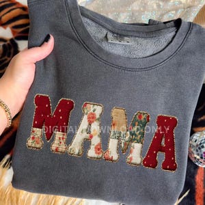 May include: A gray sweatshirt with the word "MAMA" embroidered across the front. The letters are in a patchwork style, with floral and patterned fabrics. The edges of the letters are outlined with a gold-colored thread.
