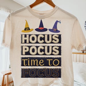 Op de afbeelding: Crèmekleurig T-shirt met drie heksenhoeden in geel, marineblauw en paars. Het shirt heeft de woorden "Hocus Pocus Time To Focus" in een gestikt ontwerp. Het shirt hangt aan een houten hanger.