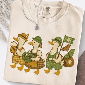 Puede incluir: Camiseta Comfort Colors color crema con un diseño caprichoso de tres patos vestidos con atuendos tradicionales alemanes. Los patos llevan cerveza, pretzels y una bandera. Un collar plateado con un colgante está sobre la camiseta.