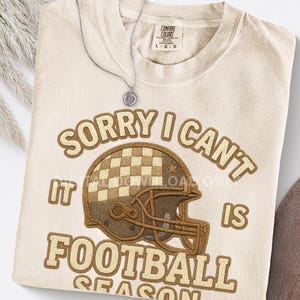 Puede incluir: Camiseta color crema con el texto "SORRY I CAN'T IT IS FOOTBALL SEASON" en letras marrones. El diseño incluye un casco de fútbol americano con un patrón de cuadros. Un collar plateado con un colgante está sobre la camiseta.