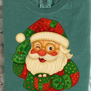 Puede incluir: Una camiseta de manga larga verde azulado con un gráfico de Papá Noel. Papá Noel guiña un ojo y sostiene una taza. El gráfico de Papá Noel tiene un diseño de patchwork con acentos rojos, verdes y blancos. La camisa está hecha de un material suave y cómodo.
