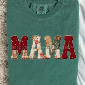 May include: A teal green sweatshirt with the word "MAMA" appliqued across the front. The letters are a mix of floral and patterned fabrics, with red polka dot fabric on the M's and A. The shirt has a crew neck and long sleeves.