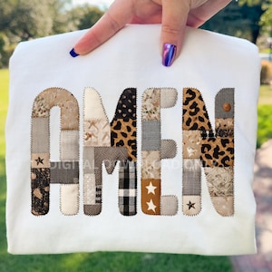 May include: White sweatshirt featuring the word "AMEN" in a patchwork design. The letters are composed of various fabric patterns, including leopard print, plaid, and stars. The sweatshirt is held up by a hand with painted nails.