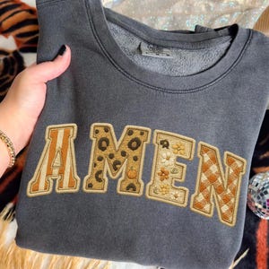 May include: A dark gray sweatshirt with the word "AMEN" embroidered across the front. The letters feature various patterns, including leopard print, floral designs, and a gingham pattern. The sweatshirt is folded and held by a hand.