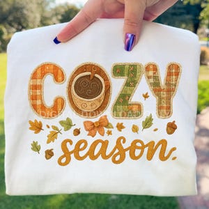 Może przedstawiać: Biała bluza z haftowanym napisem "COZY season" w jesiennych kolorach. Litery "COZY" mają wzory w kratę i motyw filiżanki kawy. Liście i żołędzie dopełniają projekt.