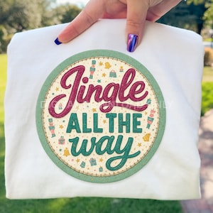 Könnte beinhalten: Weißes Sweatshirt mit einem kreisförmigen Aufdruck, der "Jingle All The Way" in Burgunderrot und Türkis zeigt. Das Design enthält festliche Symbole in einem türkisfarbenen Rahmen. Das Sweatshirt wird von einer Hand gehalten.