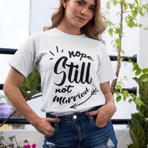 May include: A white short-sleeved t-shirt featuring black text that reads "nope Still not married" with a decorative arrow graphic pointing to the left. The shirt is paired with a dark blue denim skirt that has distressed, frayed hem details.