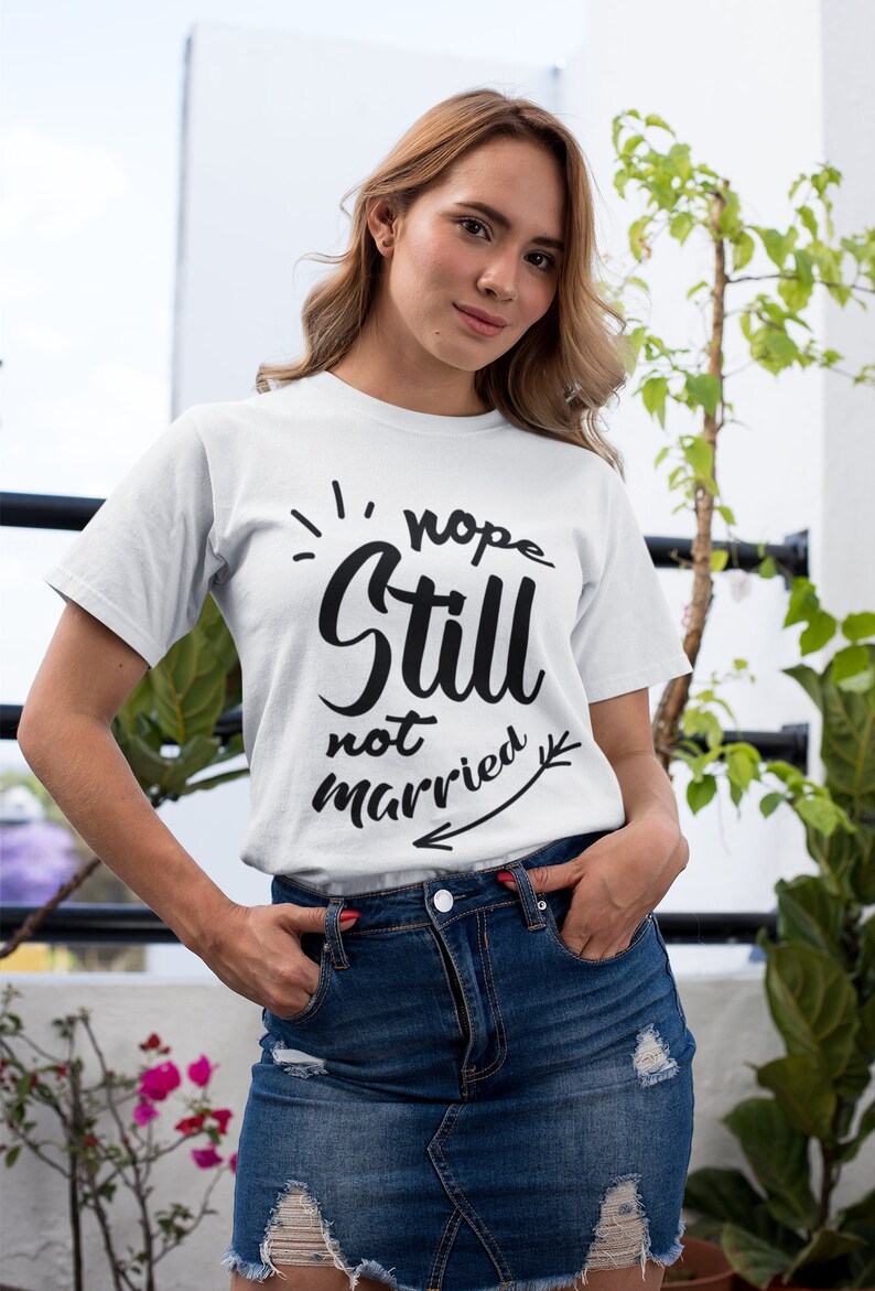 May include: A white short-sleeved t-shirt featuring black text that reads "nope Still not married" with a decorative arrow graphic pointing to the left. The shirt is paired with a dark blue denim skirt that has distressed, frayed hem details.