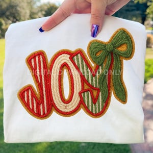 Może przedstawiać: Biała bluza z wyszytym słowem "JOY" w kolorze czerwonym i zielonym, z obramowaniem w kształcie liny. Zielona kokarda znajduje się po prawej stronie słowa. Bluza jest trzymana przez dłoń z niebieskim lakierem do paznokci.