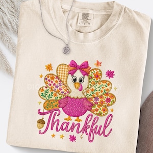 May include: Cream-colored Comfort Colors t-shirt with an embroidered turkey design. The turkey has a pink body, patterned wings, and a bow. The word "Thankful" is embroidered in pink script below the turkey. A silver necklace rests on the shirt.
