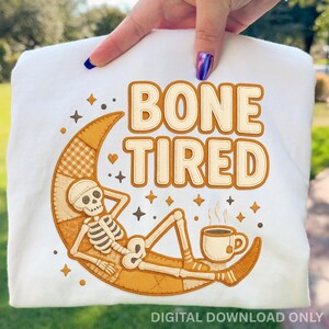 Może przedstawiać: Biała bluza z grafiką "BONE TIRED". Projekt przedstawia szkielet odpoczywający na księżycu w kształcie półksiężyca z filiżanką herbaty. Projekt jest w odcieniach beżu i brązu, z akcentami gwiazd.