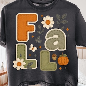 May include: Dark gray t-shirt with the word "FALL" in a patchwork design. The letters are in orange and green, with floral and leaf accents, a butterfly, and a pumpkin. The shirt is a comfortable, casual style.