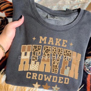 Könnte beinhalten: Dunkelgraues Sweatshirt mit dem Text "MAKE HEAVEN CROWDED" in braunen und leopardenmuster Buchstaben. Das Sweatshirt hat einen Rundhalsausschnitt und lange Ärmel. Das Design beinhaltet Sterne.