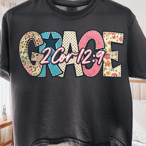 Könnte beinhalten: Dunkelgraues T-Shirt mit dem Wort "GRACE" in einem farbenfrohen Patchwork-Design. Die Buchstaben sind mit Blumen-, Leopardenmuster und Sternmustern verziert. Der Text "2 Cor 12:9" befindet sich in der Mitte des Wortes "GRACE".