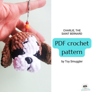 May include: A crocheted Saint Bernard dog keychain with brown, white, and black fur. The keychain has a red and orange bead charm. The text "Charlie, the Saint Bernard PDF crochet pattern by Toy Smuggler" is visible in the image.