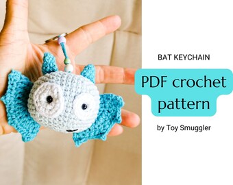 Puede incluir: Un llavero de murciélago tejido en azul y blanco con grandes ojos blancos. El llavero se sostiene en una mano y el texto "BAT KEYCHAIN PDF crochet pattern by Toy Smuggler" es visible en el fondo.