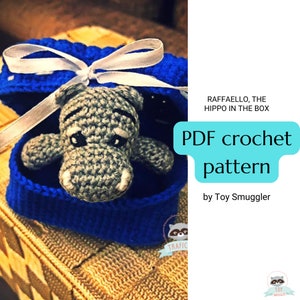 May include: A crocheted gray hippo toy inside a blue box tied with a white ribbon. The text "PDF crochet pattern" is displayed on the right, along with the text "RAFFAELLO, THE HIPPO IN THE BOX".