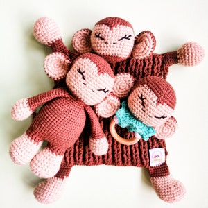 Op de afbeelding: Een bruin gehaakt aapje baby speelgoed met een rammelaar en twee kleinere aapjes eraan vast. De aapjes hebben gesloten ogen en roze wangen.