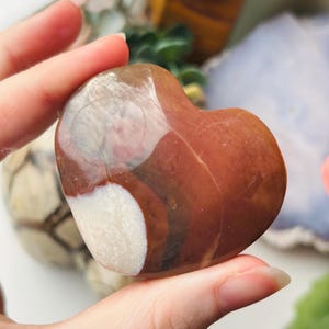 May include: A heart-shaped stone with a polished finish. The stone displays a blend of colours, including browns, white, and a touch of pink. The heart is held in a hand, highlighting its smooth texture and natural beauty.