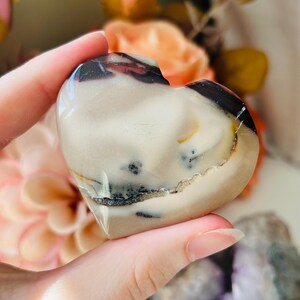 May include: A heart-shaped stone with a polished finish. The stone displays a blend of cream, brown, and black hues, with a natural pattern. The heart is held, highlighting its shape and texture. The stone is likely a decorative item.