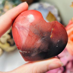 May include: A heart-shaped stone with a polished surface. The stone features a blend of red, maroon, and black hues, with natural veining patterns. The heart is held in a hand, showcasing its smooth texture and unique colour variations.