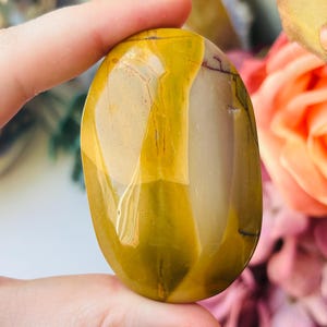 May include: A polished, oval-shaped stone with a mix of colours. The stone features a blend of yellow, cream, and brown hues, with natural veining patterns. The smooth surface reflects light, highlighting the stone's unique characteristics.
