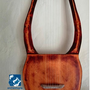 May include: A wooden lyre with a rounded body and curved arms. The instrument has strings stretched across the body and a bridge. The wood has a reddish-brown stain with darker accents. The text "Archeo Liuteria" is visible.