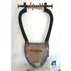 Puede incluir: Una lira antigua con un cuerpo metálico desgastado y brazos negros curvados. El instrumento tiene cuerdas y pequeñas clavijas de afinación de madera. La lira está montada en una pared blanca. La imagen incluye el texto "Archeo Liuteria".