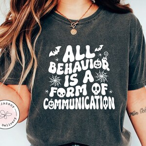 Puede incluir: Una camiseta negra con un gráfico blanco que dice "Todo comportamiento es una forma de comunicación" con ilustraciones de temática de Halloween.