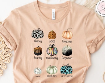 May include: A light brown t-shirt with a graphic of nine pumpkins, each with a different pattern. The pumpkins are labeled with words related to speech therapy, including "fluency", "voice", "language", "hearing", "swallowing", "cognition", "social", "modalities", and "articulation".