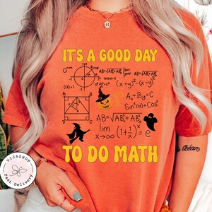 Pode incluir: Camiseta laranja com o texto "It's a good day to do math" em letras amarelas. A camiseta apresenta uma variedade de equações e símbolos matemáticos, incluindo um gráfico, um triângulo e uma equação limite. Também há ilustrações com tema de Halloween, incluindo uma abóbora, uma bruxa e um fantasma.