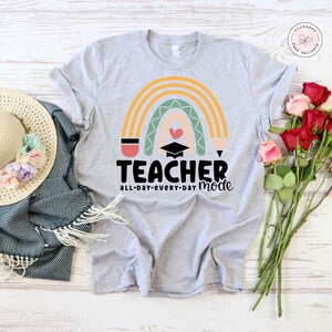 Pu&ograve; includere: Una maglietta grigia con un design a arcobaleno e il testo "TEACHER all-day-every-day mode". L'arcobaleno &egrave; composto da strisce gialle, arancioni, verdi e rosa. Al centro dell'arcobaleno c'&egrave; un cappello da laurea e un cuore.