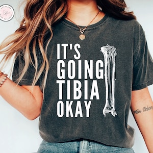 May include: A dark gray t-shirt with the text "It's Going Tibia Okay" in white. A white anatomical illustration of a lower leg bone is to the right of the text.